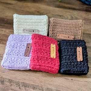 Op de afbeelding: Vijf handgemaakte gehaakte hoezen in crème, bruin, lavendel, rood en zwart. Elke hoes heeft een rechthoekige vorm en een klein leren label met de tekst "Hand Made". De hoezen zijn gerangschikt op een houten oppervlak.