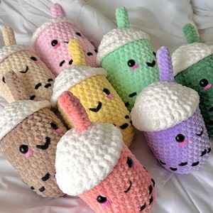 May include: A collection of eight crocheted plush bubble tea toys in various colours, including pink, green, yellow, brown, purple, and white. Each toy has a smiling face with black eyes and a white crocheted lid.