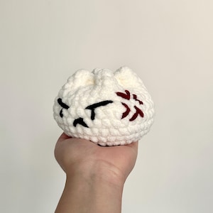 Peut inclure: Une pochette blanche crochetée avec des détails brodés noirs et rouges. La pochette présente un motif de visage grincheux avec des yeux noirs et un élément floral rouge stylisé. L'article fait main est tenu dans une main sur un fond uni.