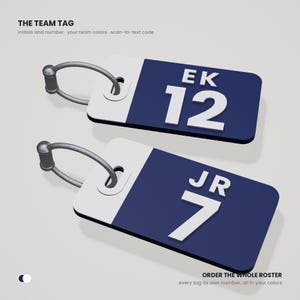 May include: Two rectangular luggage tags with a white and dark blue color scheme. Each tag features white raised initials and a number, specifically EK 12 and JR 7. A grey cord loop is attached to each tag. Text reads THE TEAM TAG and ORDER THE WHOLE ROSTER.