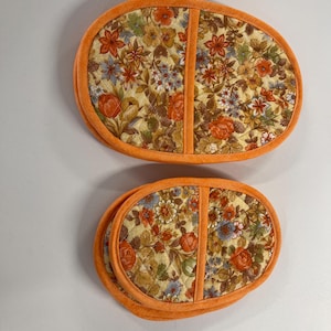 May include: Two oval-shaped quilted pot holders. The floral fabric features orange, red, yellow, and blue flowers on a cream background. The edges are trimmed with orange fabric. The pot holders are designed to protect surfaces from heat.