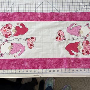 May include: A white table runner with a pink border features four gnomes holding heart-shaped balloons. The gnomes are wearing pink and red hats and have white beards. The balloons are decorated with pink and white hearts.