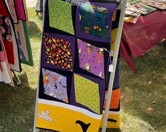 Könnte beinhalten: Eine Halloween-Steppdecke mit einem gelben, weißen und schwarzen Rand. Die Steppdecke besteht aus verschiedenen Quadraten mit verschiedenen Halloween-Motiven, darunter Kürbisse, Fledermäuse, Geister und Mais.