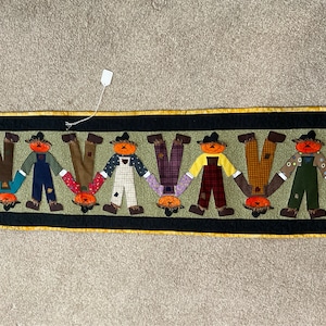 Può includere: Un arazzo da parete trapuntato con una fila di otto spaventapasseri capovolti con teste di zucca. Ogni spaventapasseri è vestito con abiti patchwork diversi, tra cui salopette, camicie a quadri e pantaloni a righe. Gli spaventapasseri sono disposti in fila su uno sfondo nero con un bordo giallo.