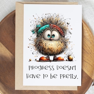 May include: A greeting card featuring a fluffy, round, brown creature with large blue eyes, wearing a teal and red patterned headband and orange shoes. The text below reads: PROGRESS DOESN'T HAVE TO BE PRETTY. The card is set against a white background.