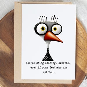 May include: Greeting card with a whimsical bird illustration. The bird has large, expressive eyes, a red beak, and a black and white polka-dot pattern on its neck. The text reads, "You're doing amazing, sweetie... even if your feathers are ruffled."