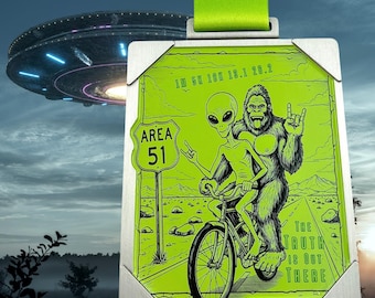 May include: A rectangular medal with a neon green graphic of an alien and Bigfoot riding a bicycle on a road, with the text "The Truth Is Out There." The medal has the text "1M 5K 10K 13.1 26.2" and "Area 51" on it. A green ribbon is attached.