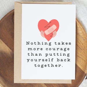 May include: A greeting card with a red heart graphic, a bandage, and the text "Nothing takes more courage than putting yourself back together." The card is on a wooden tray with a brown envelope.