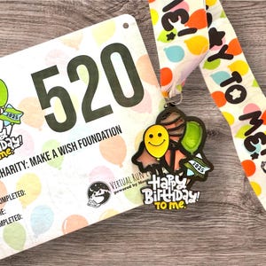 May include: A white and pink running bib with the number 520 and the text "Charity: Make a Wish Foundation". A colorful medal with the text "Happy Birthday! To me." and a ribbon with the text "Hey to Me!" are also shown.