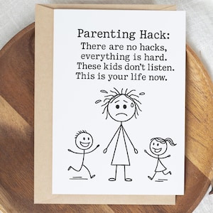 May include: A white greeting card with the text "Parenting Hack: There are no hacks, everything is hard. These kids don't listen. This is your life now." features a stick figure parent with two stick figure children. The card is inside a tan envelope.