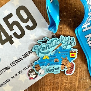 May include: A blue ribbon with white stars and the text "Kentucky" on it. A blue and white medal with a map of Kentucky and the text "Kentucky's" on it. The medal is attached to the ribbon. The medal features a horse, a baseball bat, a banjo, and a bucket of fried chicken.