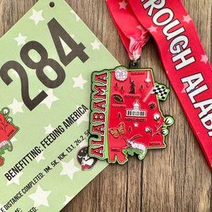 May include: A red, white, and blue ribbon with the text "ROUGH ALABAMA" printed on it. A metal medal in the shape of the state of Alabama with a red, white, and blue design. The medal has the word "ALABAMA" printed on it. A green and white card with the number "284" printed on it. The card also has the text "BENEFITTING: FEEDING AMERICA" and "DISTANCE COMPLETED: 1M, 5K, 10K, 13.1, 26.2, 52"