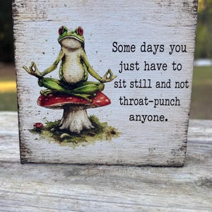 May include: A square wooden sign with a distressed white finish featuring a green frog with red eyes sitting in a meditative pose on a red mushroom. The text reads: Some days you just have to sit still and not throat-punch anyone.