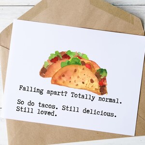 May include: A white greeting card with a watercolor illustration of two tacos, filled with meat, lettuce, and tomatoes. The card reads, "Falling apart? Totally normal. So do tacos. Still delicious. Still loved."