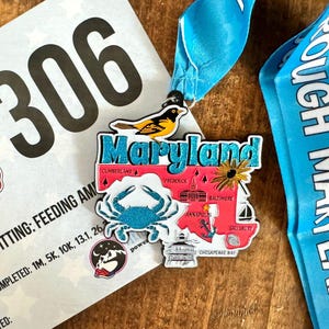 May include: A Maryland Tough Mudder race medal with a blue ribbon. The medal features a map of Maryland with a crab, a bird, and the word "Maryland". A race bib with the number 306 is also visible.
