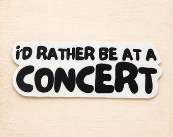 May include: A white rectangular sticker with rounded edges featuring the black text I'D RATHER BE AT A CONCERT in a bold, rounded, bubble-style font. The sticker has a clean, die-cut appearance with high contrast between the black lettering and white background.
