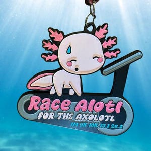 May include: A pink and white axolotl medal with a treadmill design. The axolotl has pink gills and is on a black treadmill. The medal reads "Race Axolotl for the Axolotl" with race distances.