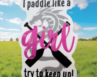 May include: A white sticker with the text "I paddle like a girl try to keep up!" in black and pink. The sticker features a gray dragon design and crossed black paddles. The background is a field and blue sky.