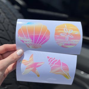 May include: Four iridescent seashell-themed stickers. The stickers feature a seashell, sand dollar, starfish, and conch shell. The colors transition from pink to orange and yellow, with a hint of blue.