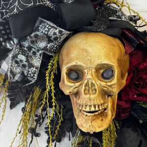 May include: A Halloween wreath featuring a realistic-looking skull with black and silver ribbon, black and white checkered ribbon, and red roses.
