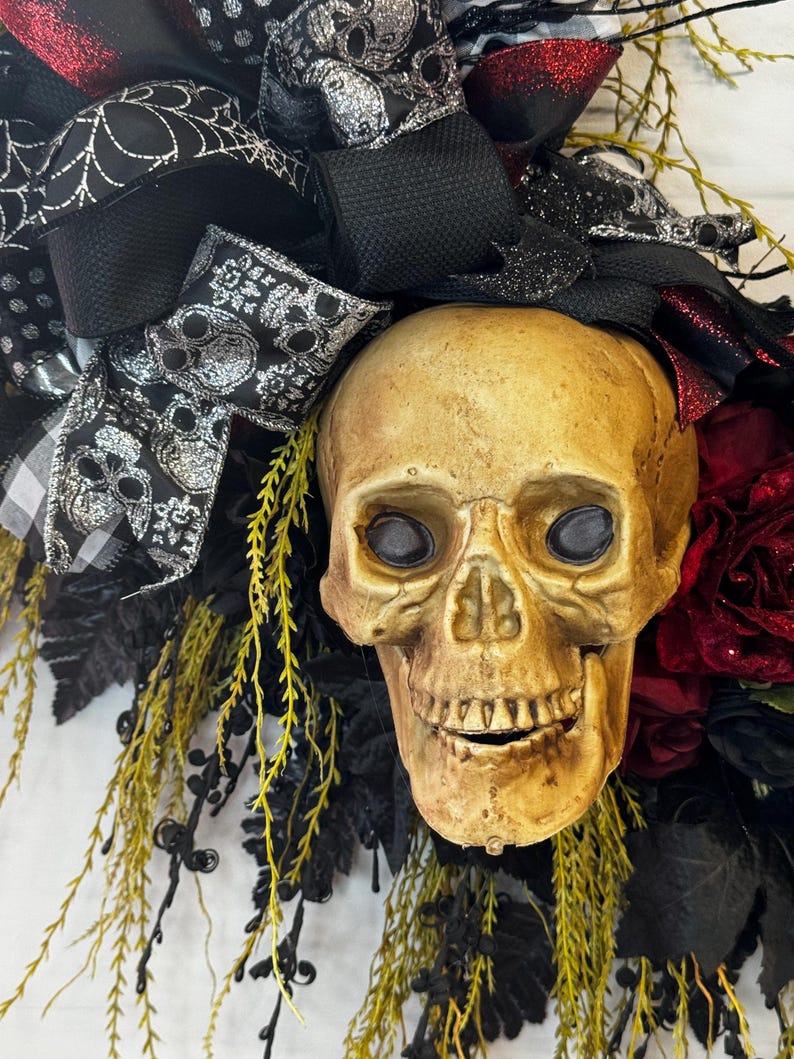 May include: A Halloween wreath featuring a realistic-looking skull with black and silver ribbon, black and white checkered ribbon, and red roses.