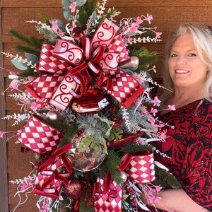 May include: A festive Christmas decoration featuring a mix of evergreen branches, red and white diamond patterned ribbons, and velvet accents. The arrangement includes pink floral accents and decorative ornaments.