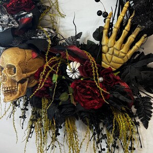 May include: A Halloween wreath with a skull, a skeleton hand, red roses, black and white ribbon, and green foliage.