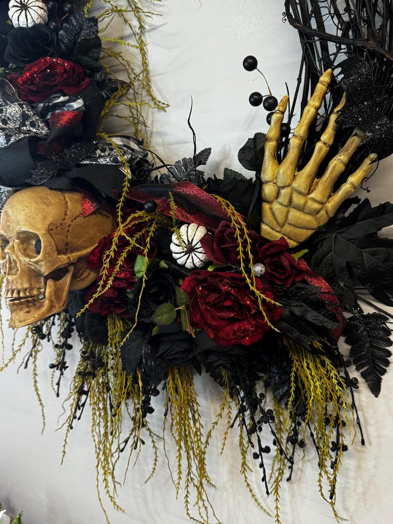 May include: A Halloween wreath with a skull, a skeleton hand, red roses, black and white ribbon, and green foliage.