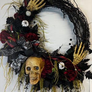 May include: A black twig wreath decorated with black and red roses, a skull, and skeletal hands. The wreath is accented with black and white ribbon and spiderwebs.