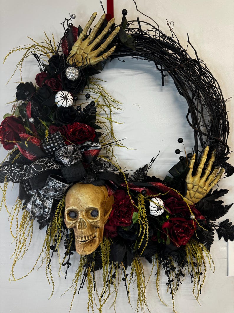 May include: A black twig wreath decorated with black and red roses, a skull, and skeletal hands. The wreath is accented with black and white ribbon and spiderwebs.