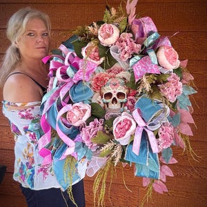 May include: A pink and blue floral wreath with a sugar skull in the center. The wreath is decorated with pink and blue ribbons and flowers.