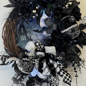 May include: A Halloween wreath with a grapevine base, adorned with black and white ribbons, spiderwebs, and black flowers. A silhouette of a haunted house and bats is visible in the center. The wreath is decorated with small lights.