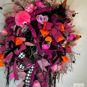 May include: A pink and black Halloween wreath with a spider, witch hat, and skeleton bones. The wreath is decorated with pink, orange, and black ribbons and flowers.