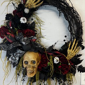 May include: A black twig wreath decorated with red and black roses, a skull, and skeletal hands. The wreath is accented with white and black spiderwebs, black berries, and white pumpkins.