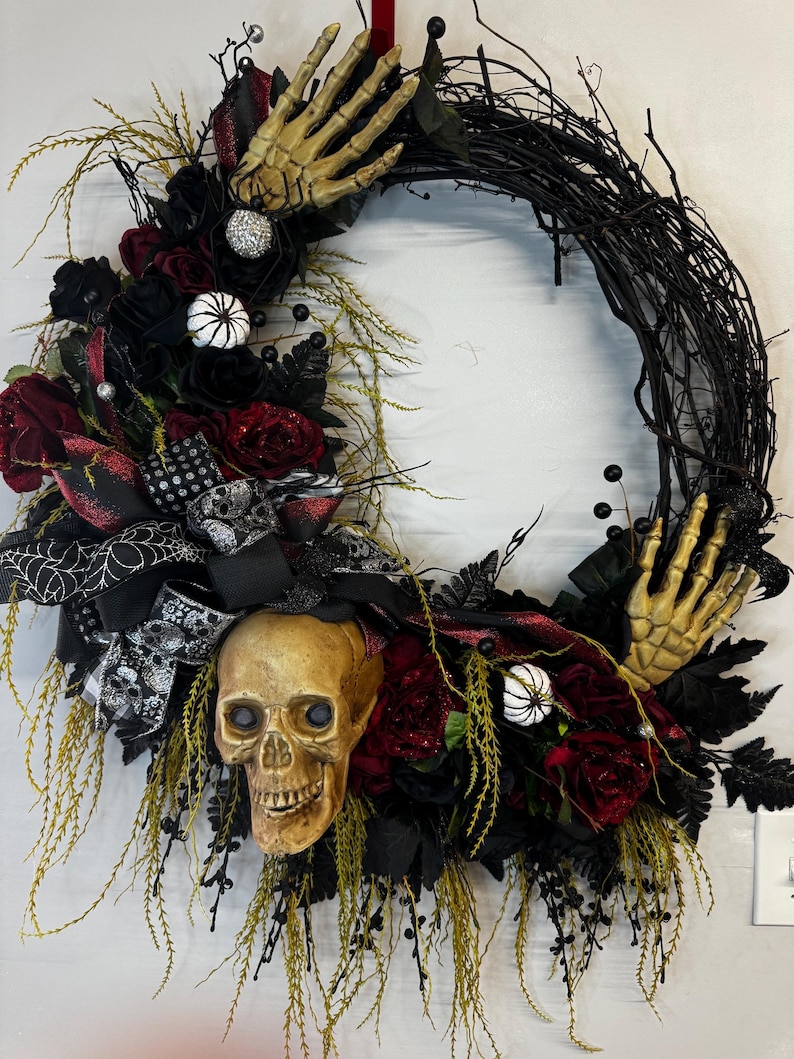 May include: A black twig wreath decorated with red and black roses, a skull, and skeletal hands. The wreath is accented with white and black spiderwebs, black berries, and white pumpkins.