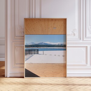 May include: A framed photograph of a snowy landscape with a lake and mountains in the background. The photo is framed in a light wood frame and is hanging on a white wall.