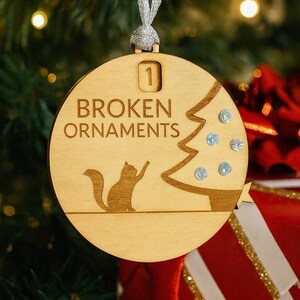 May include: Wooden Christmas ornament with the text "BROKEN ORNAMENTS" and a cat silhouette reaching towards a Christmas tree. The ornament has a silver ribbon for hanging and is decorated with small, sparkling embellishments.