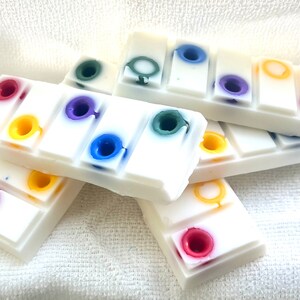 Op de afbeelding: Een stapel witte wasmelts met kleurrijke cirkels erop. De wasmelts zijn in de vorm van een staaf en hebben een glad, wit oppervlak. De kleuren van de cirkels zijn rood, geel, blauw, groen en paars.