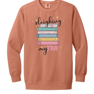 May include: A coral-colored sweatshirt with the words "sleighing my TBR" and a stack of colorful books. The design includes a pink, yellow, blue, and green book. The text is in a whimsical font with star accents.
