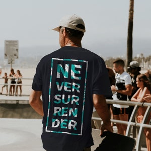 Op de afbeelding: Een zwart T-shirt met een wit rechthoek en de tekst "NEVER SURRENDER" in turquoise en witte letters.