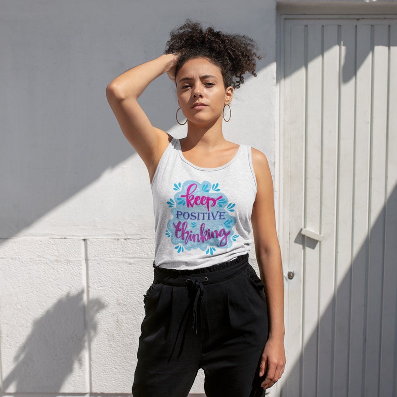 May include: A woman wearing a gray tank top with the text "keep positive thinking" in pink and blue lettering. The tank top has a watercolor-like design with blue and white clouds.