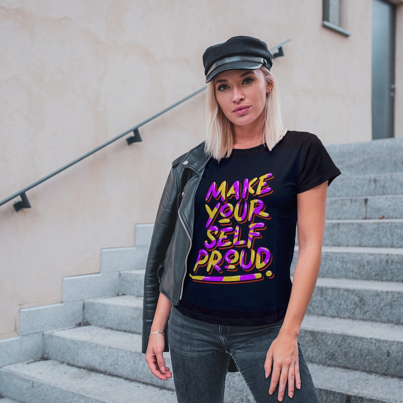 Op de afbeelding: Een vrouw draagt een zwart T-shirt met de tekst "MAKE YOUR SELF PROUD" in paarse en gele letters. Ze draagt een zwart leren jack en een zwarte platte pet. Ze staat op een betonnen trap.