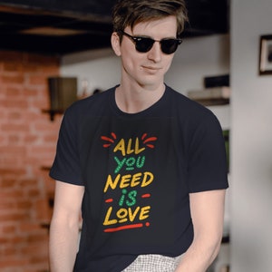 Op de afbeelding: Een donkerblauw t-shirt met een kleurrijke afbeelding met de tekst "ALL YOU NEED IS LOVE".