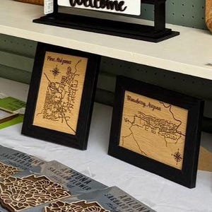 May include: Two framed wooden maps with black frames. One map is labeled "Pine, Arizona" and the other "Strawberry, Arizona." The maps are laser-cut with intricate details of the towns' layouts. Below are several Arizona Saguaro Flower cards.