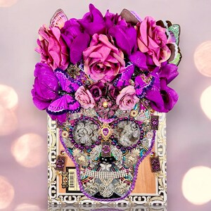 May include: A purple and pink floral arrangement with butterflies, covering a wooden box with a skull design made of rhinestones and other embellishments.