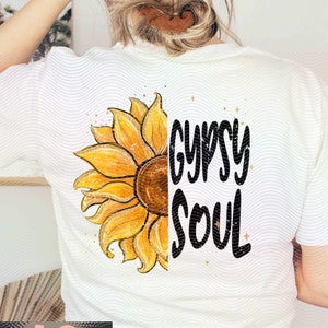 Puede incluir: Camiseta blanca con un gran diseño de girasol en el lado izquierdo y las palabras "Gypsy Soul" en escritura negra en el lado derecho. El girasol es amarillo y marrón con detalles dorados.