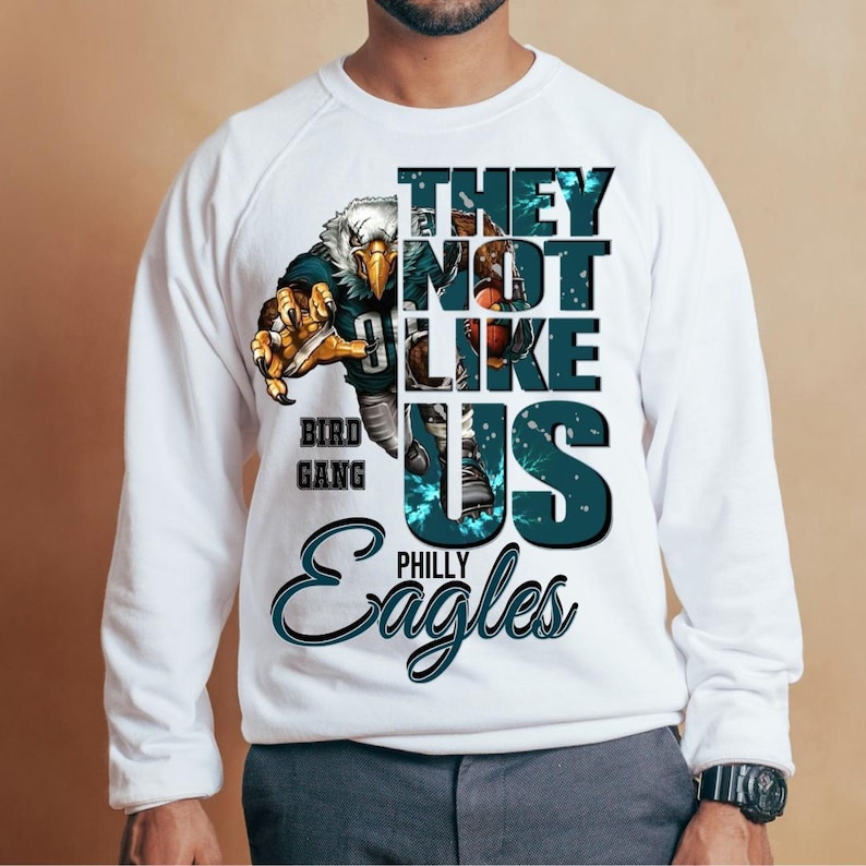 Może przedstawiać: Biała bluza z nadrukiem orła i napisem "THEY NOT LIKE US" oraz "PHILLY Eagles". Orzeł ma na sobie str&oacute;j do futbolu amerykańskiego. Na bluzie wydrukowano r&oacute;wnież słowa "BIRD GANG".