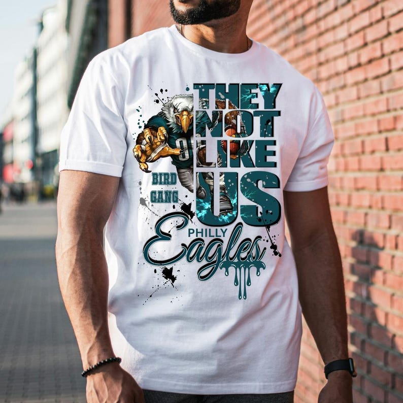 Może przedstawiać: Biała koszulka z grafiką przedstawiającą orła, napisem "THEY NOT LIKE US" i "PHILLY Eagles". Projekt zawiera kolory turkusowy, czarny i złoty, z efektem rozprysku i postarzenia.
