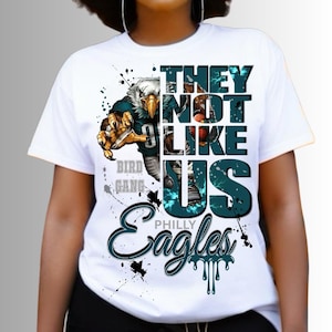 Może przedstawiać: Biała koszulka z grafiką orła futbolowego i napisem "THEY NOT LIKE US" oraz "PHILLY EAGLES". Projekt zawiera turkusowe i czarne akcenty z efektami rozprysk&oacute;w farby.