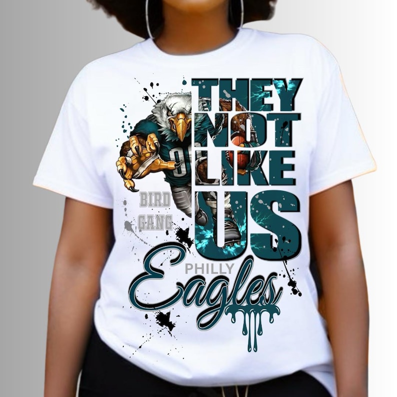 Może przedstawiać: Biała koszulka z grafiką orła futbolowego i napisem "THEY NOT LIKE US" oraz "PHILLY EAGLES". Projekt zawiera turkusowe i czarne akcenty z efektami rozprysk&oacute;w farby.